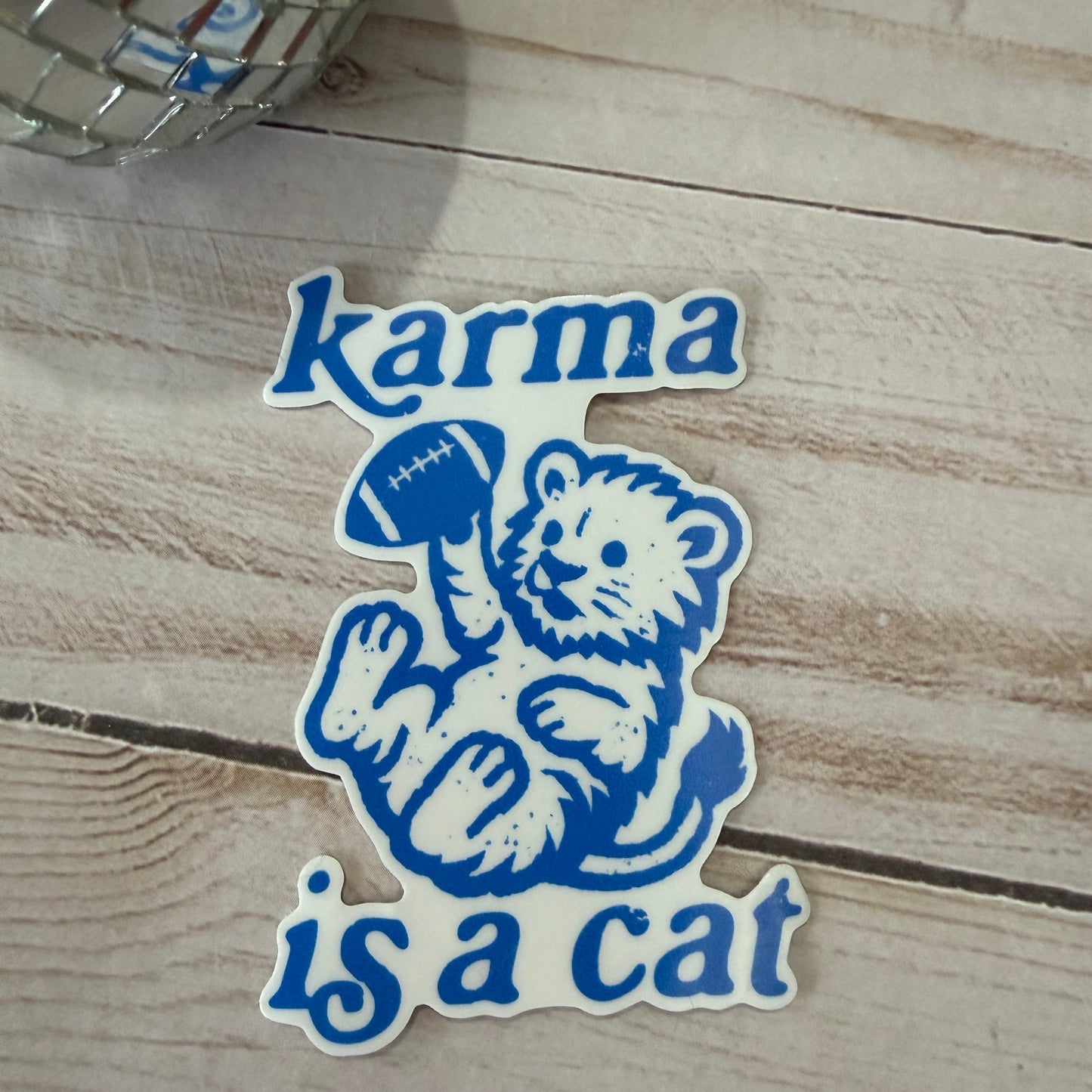 Sticker with bear holding a football and 'karma is a cat' text on a wooden surface