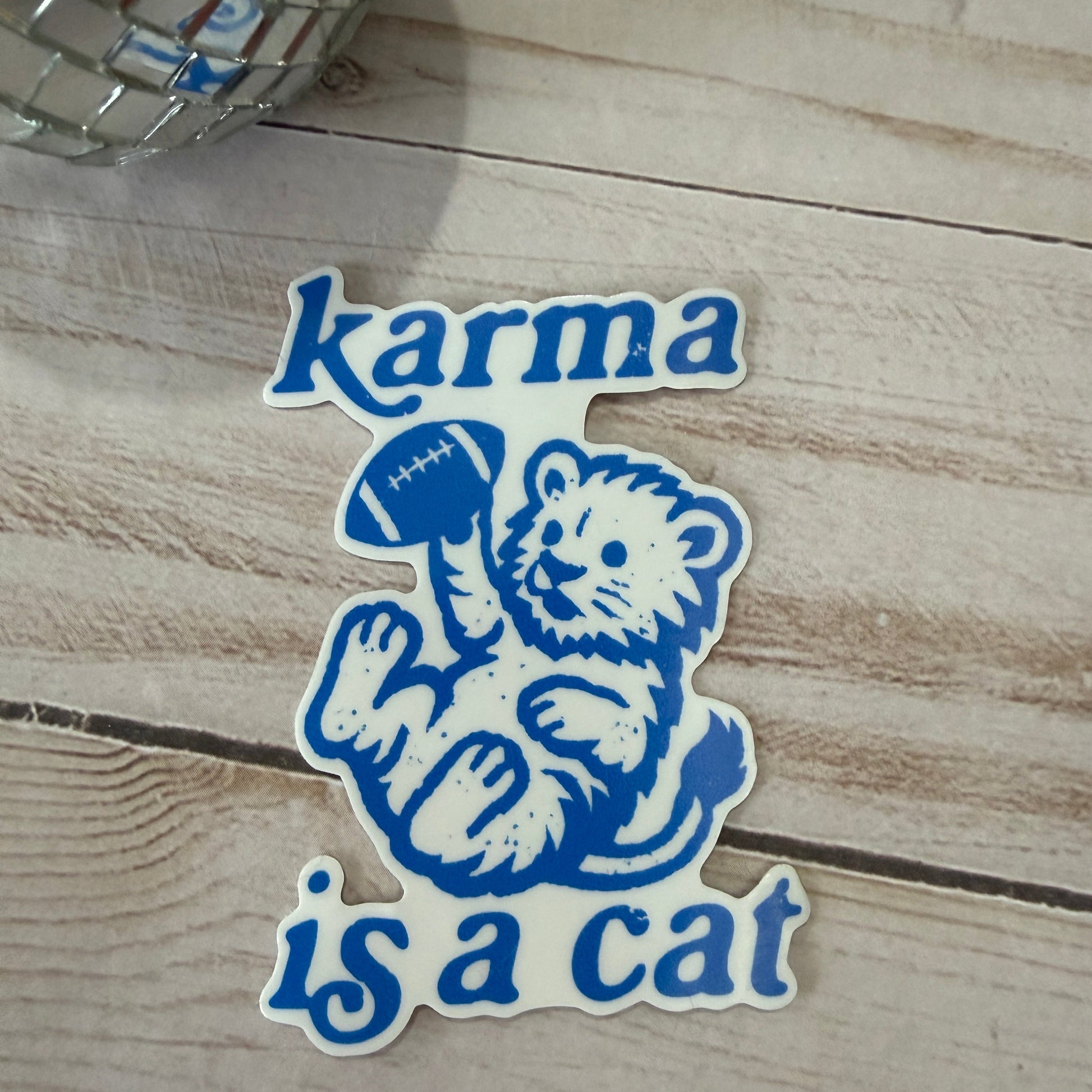 Sticker with bear holding a football and 'karma is a cat' text on a wooden surface